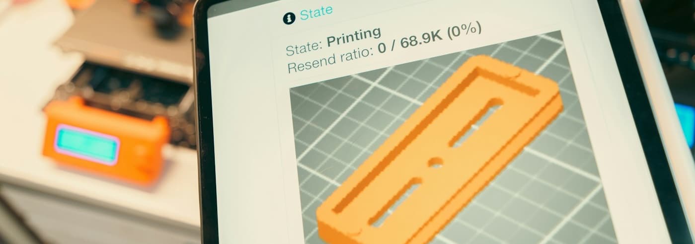 Tablet showing a 3D model of an orange part, with a 3D printer in the background